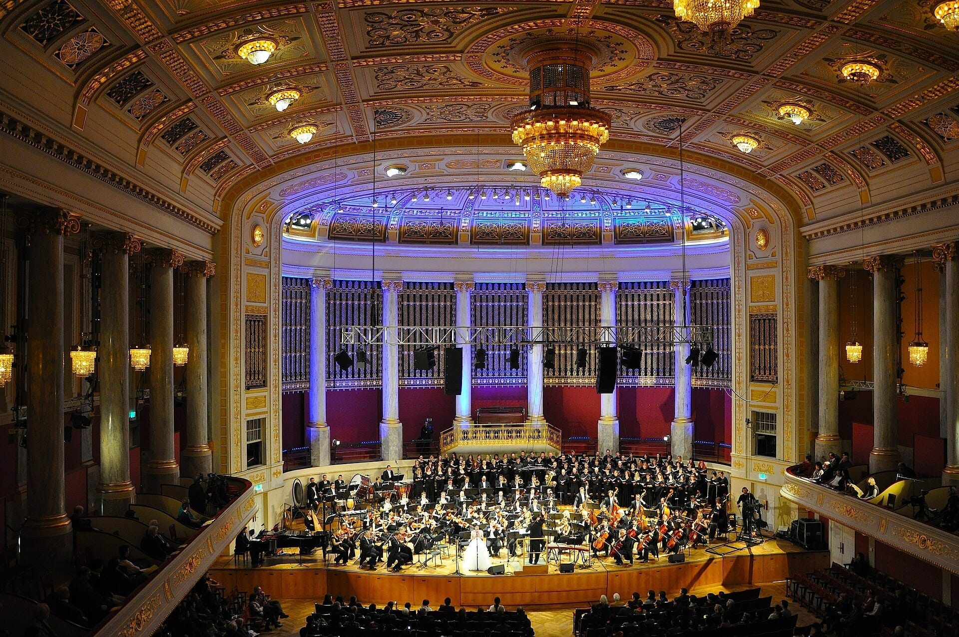 Großes Inneres des Wiener Konzerthauses mit verzierten Säulen, funkelnden Kronleuchtern und einem vollständigen Orchester, das auf der Bühne vor dem Publikum auftritt.