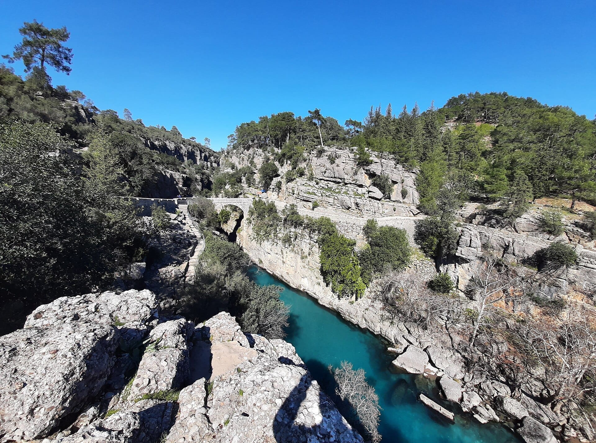 Un antico ponte in pietra attraversa il turchese fiume Köprülü in un canyon roccioso circondato da una fitta foresta verde sotto un cielo azzurro limpido.
