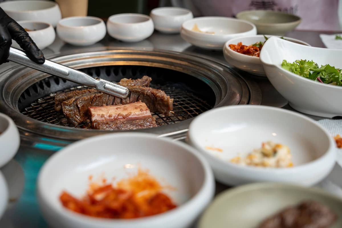 Guarnições variadas e carne marinada sendo grelhada em uma mesa de restaurante de churrasco coreano, destacando a autêntica experiência gastronômica.