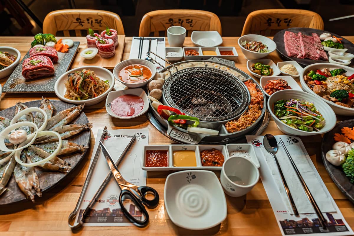 Uma mesa preparada para Korean BBQ com carne crua, acompanhamentos banchan e uma grelha embutida na mesa pronta para cozinhar.