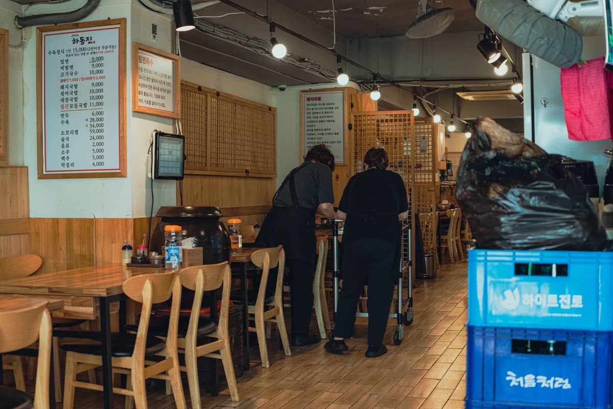 Dois clientes dentro de um restaurante coreano em Seul, com um cardápio visível na parede, mesas de madeira e uma configuração típica de jantar BBQ.