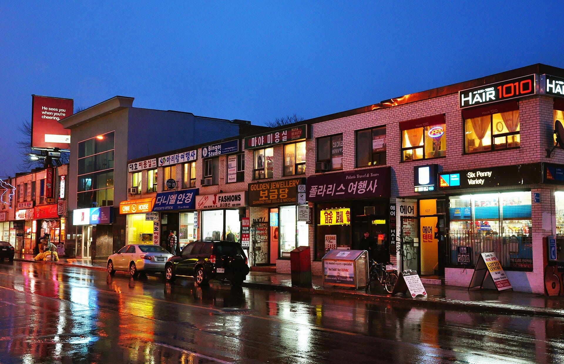 ร้านค้าที่มีป้ายภาษาเกาหลีและไฟนีออนเรียงรายตาม Bloor Street ที่เปียกและสะท้อนแสงในย่าน Koreatown ของเมือง Toronto ยามพลบค่ำ