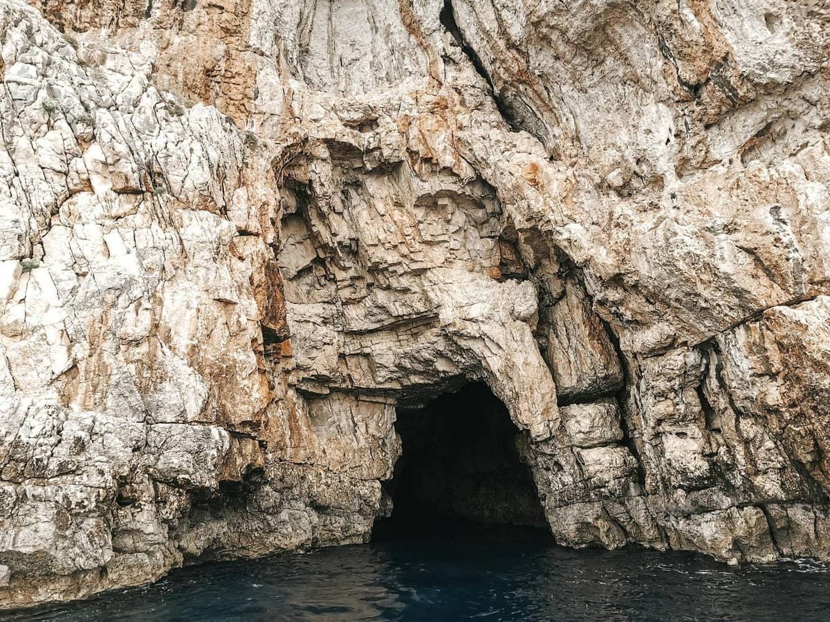 Entrance to a sea cave set in rugged limestone cliffs above deep blue water on a sunny day.