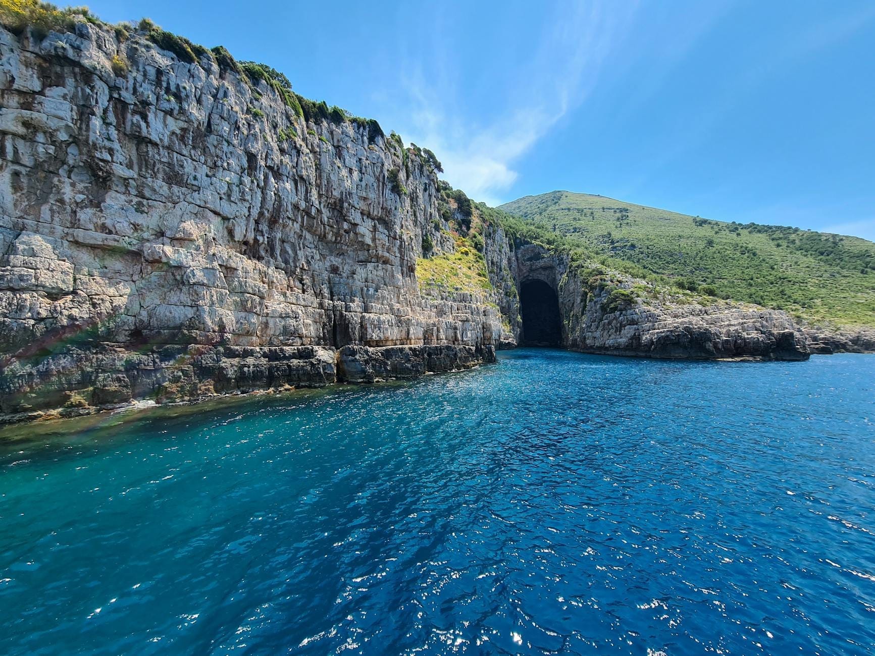Penhascos rochosos e a entrada para a Gruta Azul perto de Kotor, Montenegro, rodeados por água azul cristalina e vegetação exuberante sob um céu brilhante.