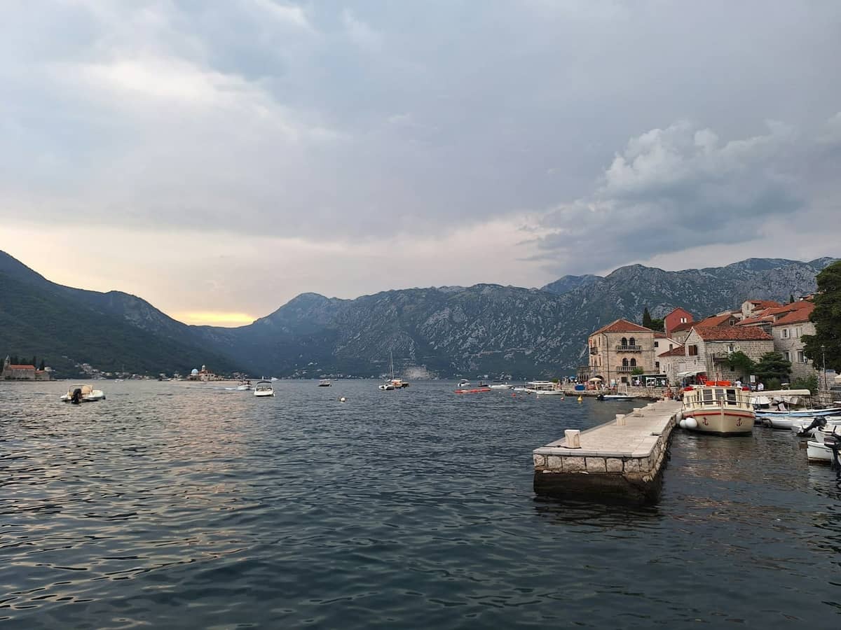 Scenic view of Kotor Bay waterfront with small boats, stone dock, and historic buildings backed by mountains at dusk.