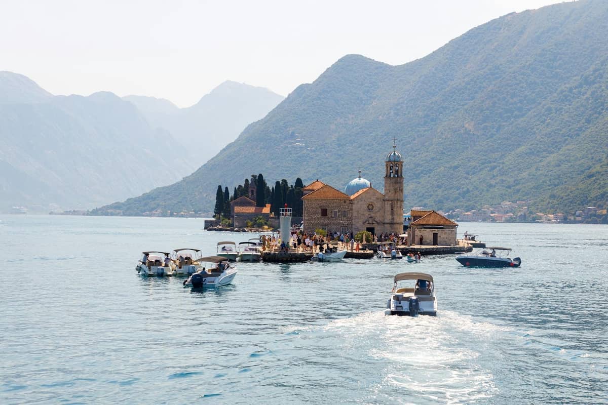 Several boats approach Our Lady of the Rocks and Saint George islands in the Bay of Kotor, set against towering green mountains.