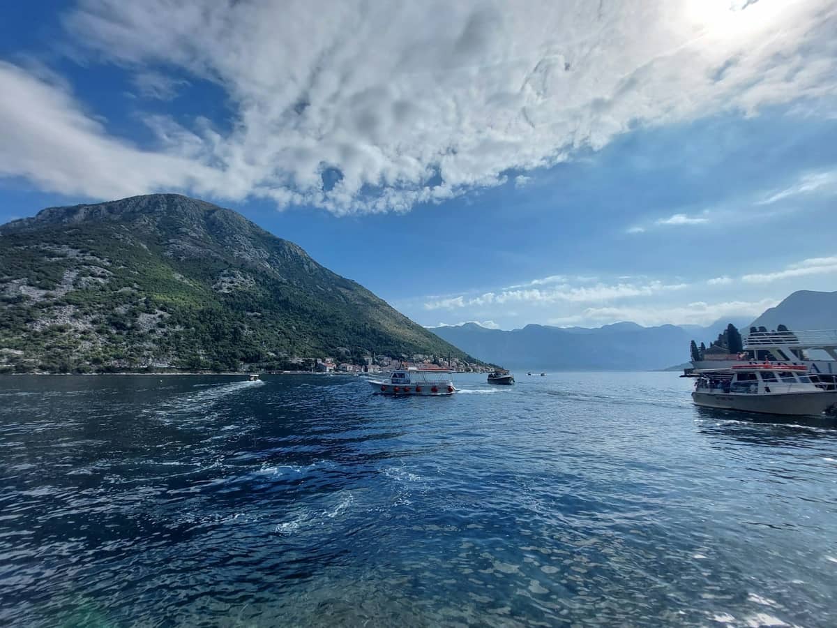 Wide view of coastal Kotor with boats on clear blue water under a sunny sky, mountains in the distance, and vibrant sea reflections.