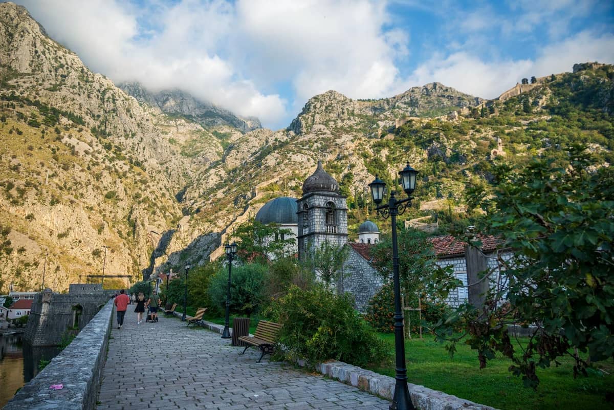 Pedestrian walkway in Kotor with people walking towards mountains, benches, and greenery alongside a stone path.