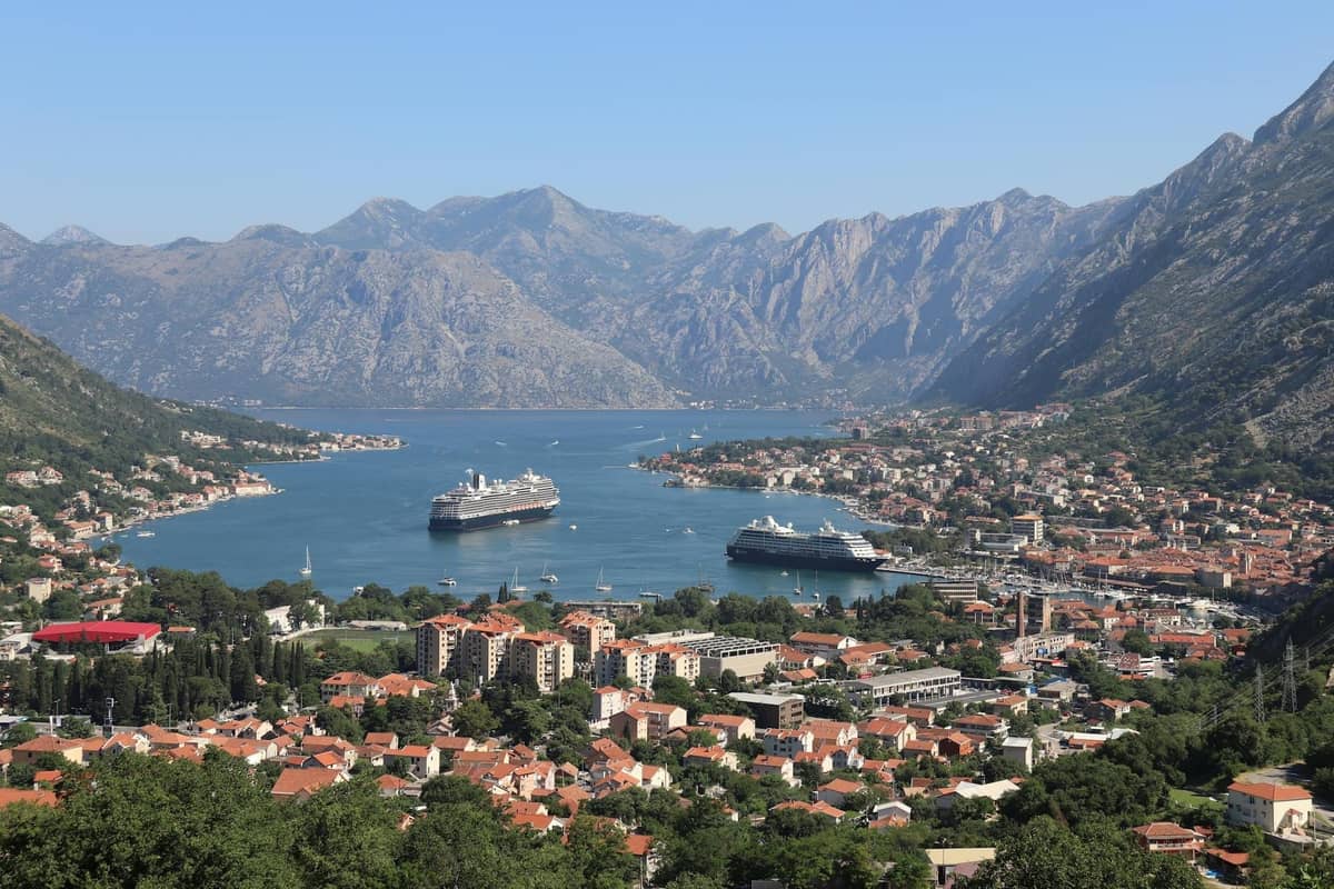 Sunny, panoramic view of Kotor Bay with cruise ships docked and clear weather over the town and mountains.