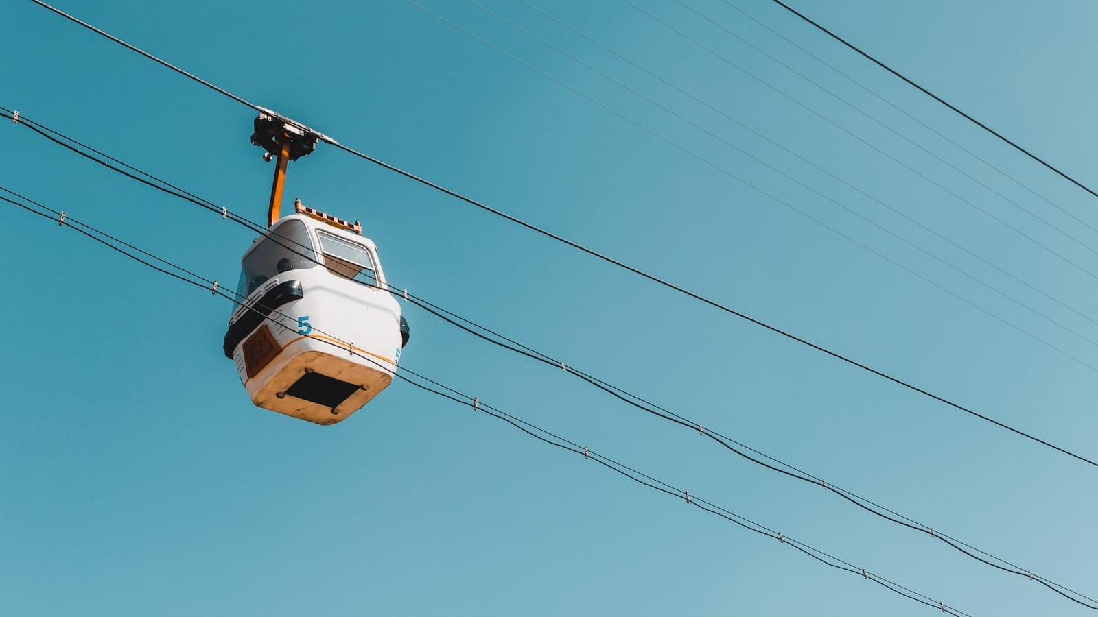 Cabine branca do teleférico de Kotor percorrendo os cabos contra um céu azul claro, fotografada de baixo em um dia ensolarado.