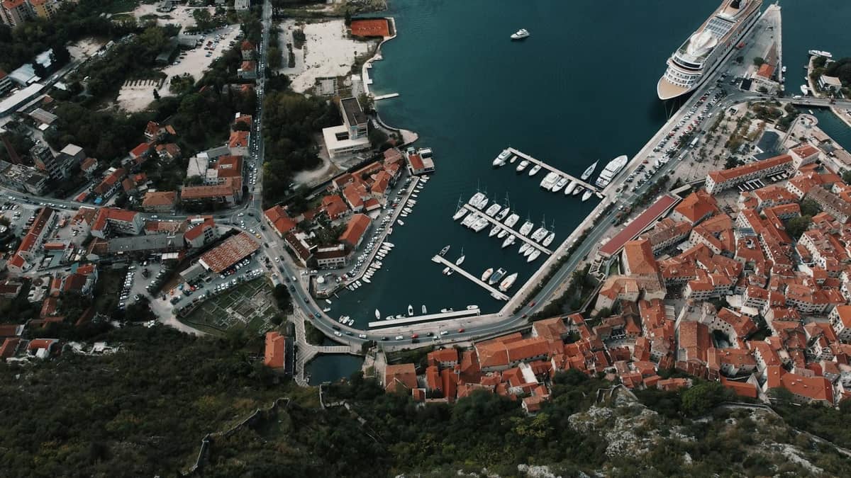 Aerial view of Kotor's cruise port with a large cruise ship docked beside the Old Town and marina, surrounded by red-roofed buildings.
