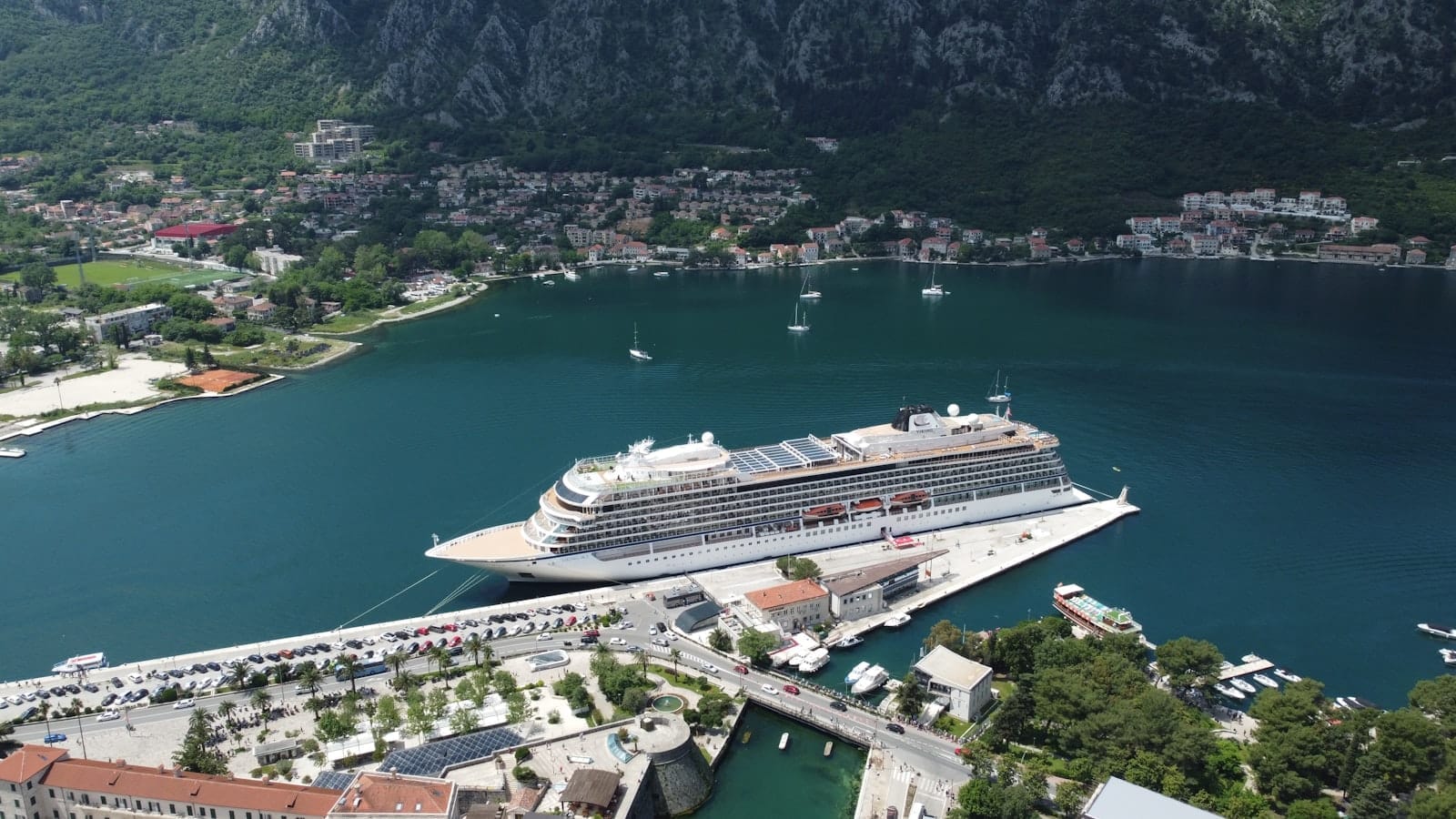 Vista aérea de um grande navio de cruzeiro atracado no Porto de Cruzeiros de Kotor, rodeado por colinas verdes, edifícios à beira-mar e águas azuis calmas.