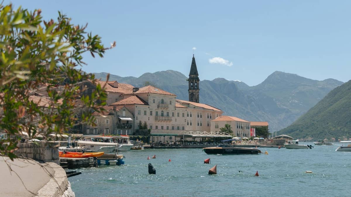 A picturesque waterfront village with stone buildings, a tall bell tower, small boats, and dramatic mountains rising in the background.