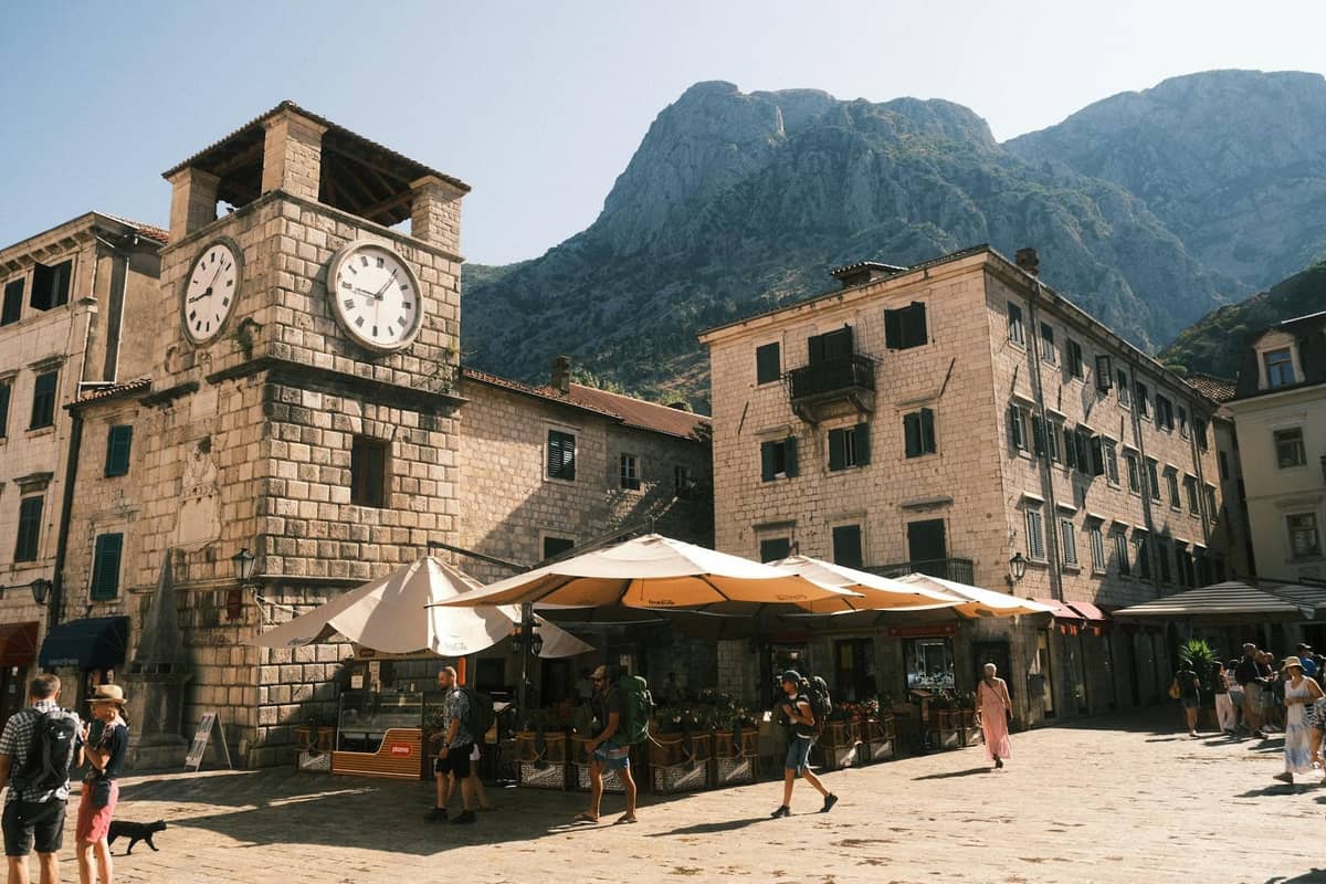 Lively square in Kotor Old Town with the famous clock tower, stone buildings, and mountains rising in the background.