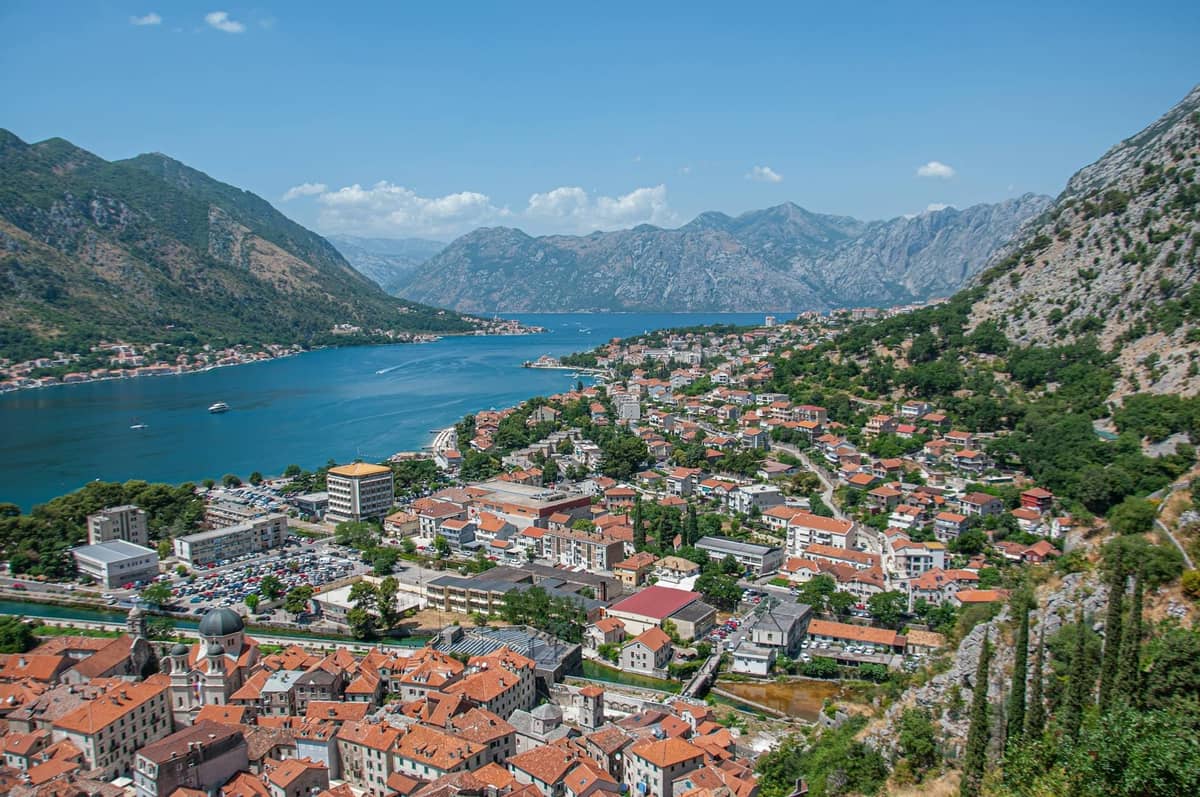 Daytime view over Kotor with red rooftops, bay waters, and surrounding mountains under a clear blue sky.