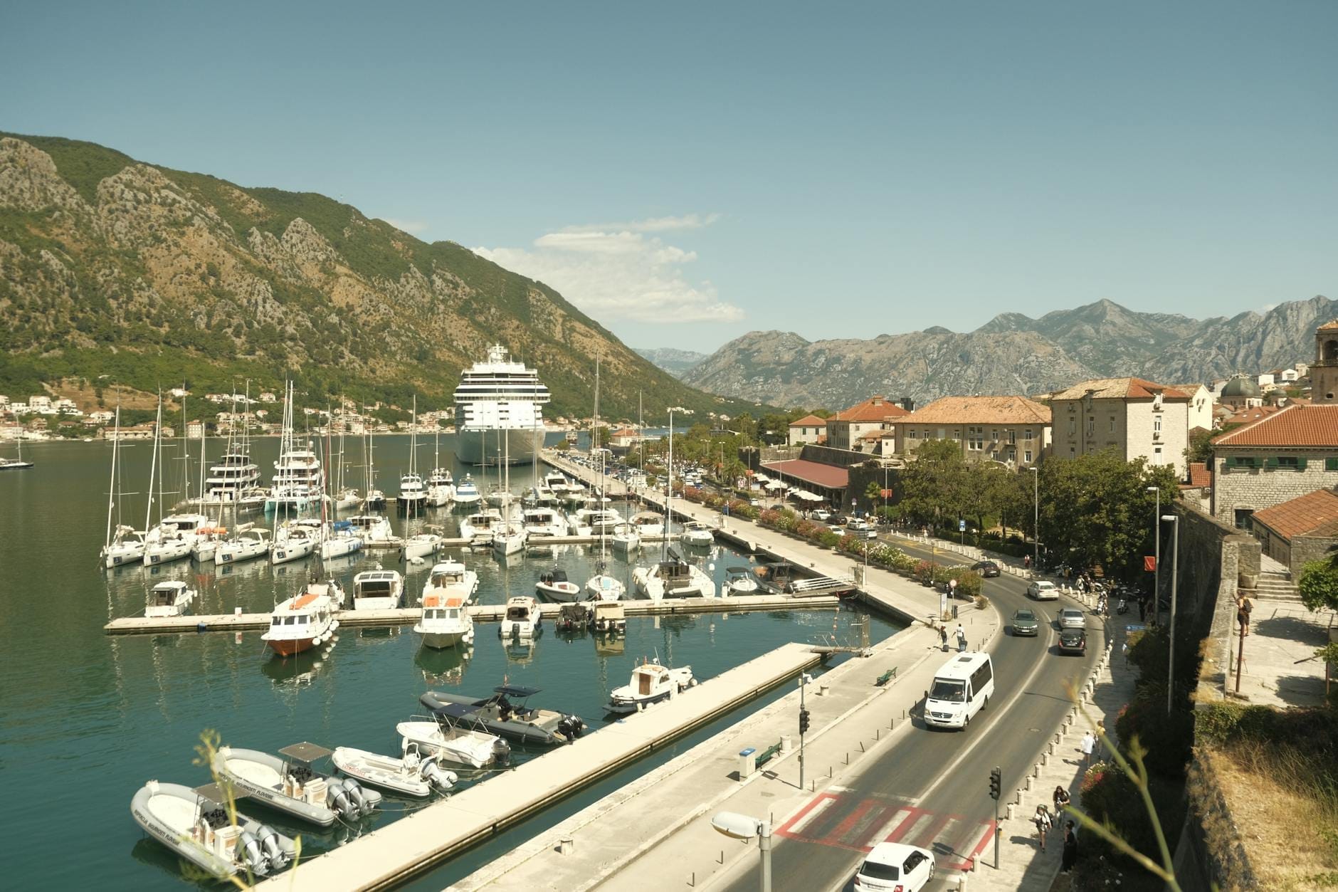 Vista della marina di Kotor con barche e yacht ormeggiati lungo il lungomare, le mura della Città Vecchia, edifici dai tetti rossi e montagne sullo sfondo sotto un cielo sereno.