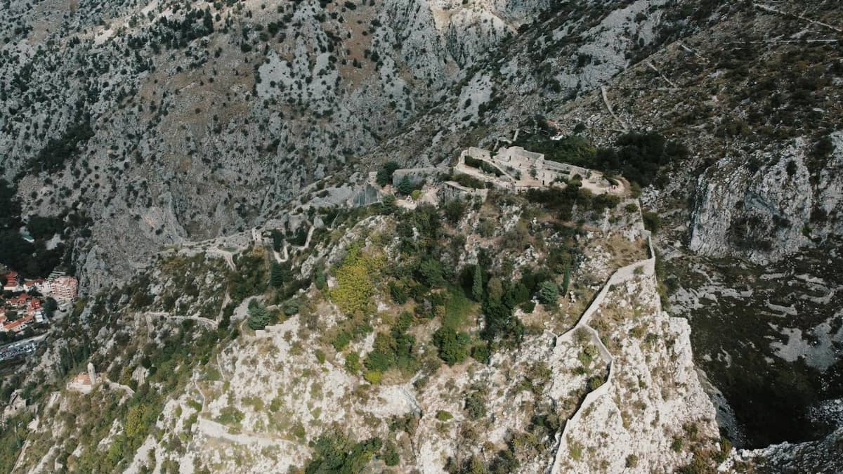 Aerial view of Kotor’s city walls winding up the steep mountainside, showing fortifications and stone steps amidst rocky terrain.