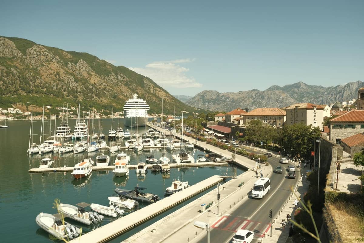 View of Kotor waterfront promenade with marina, boats, mountains and old town buildings along a flat walkway outside the city walls.