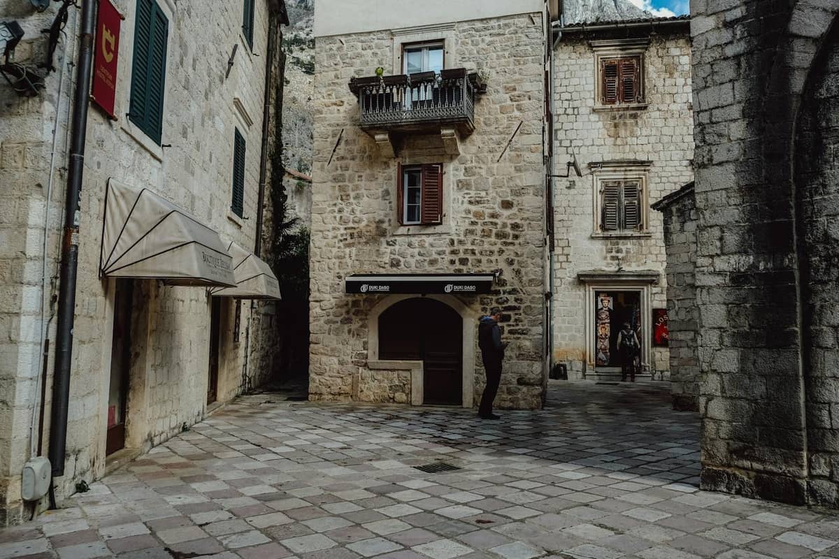 Quiet stone courtyard in Kotor old town with narrow alleys, historic stone buildings, and a few people, showing hidden residential character.