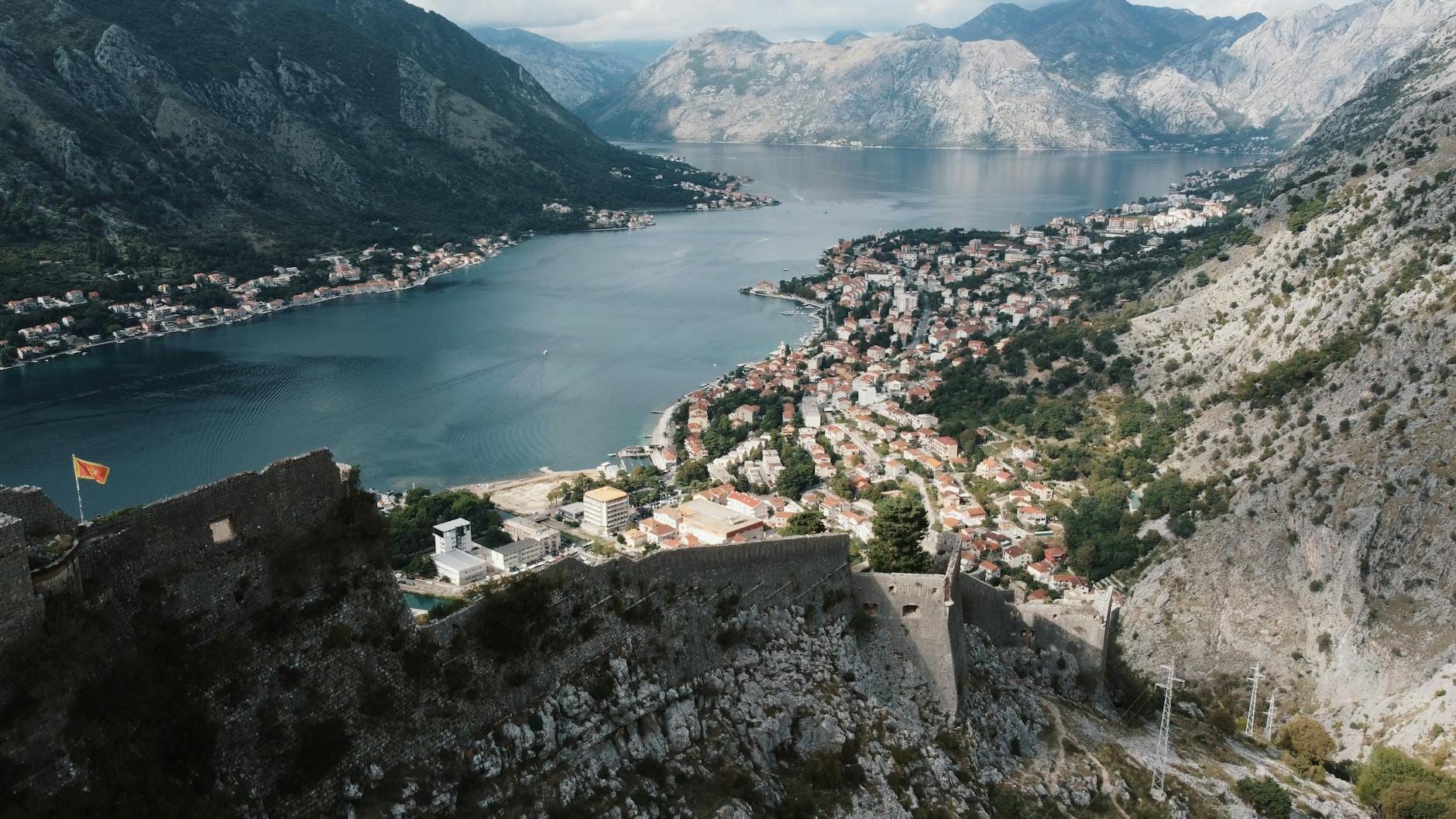 A stunning panoramic view over the Bay of Kotor, showing the walled old town below, surrounded by dramatic mountains and blue water under a partly cloudy sky.