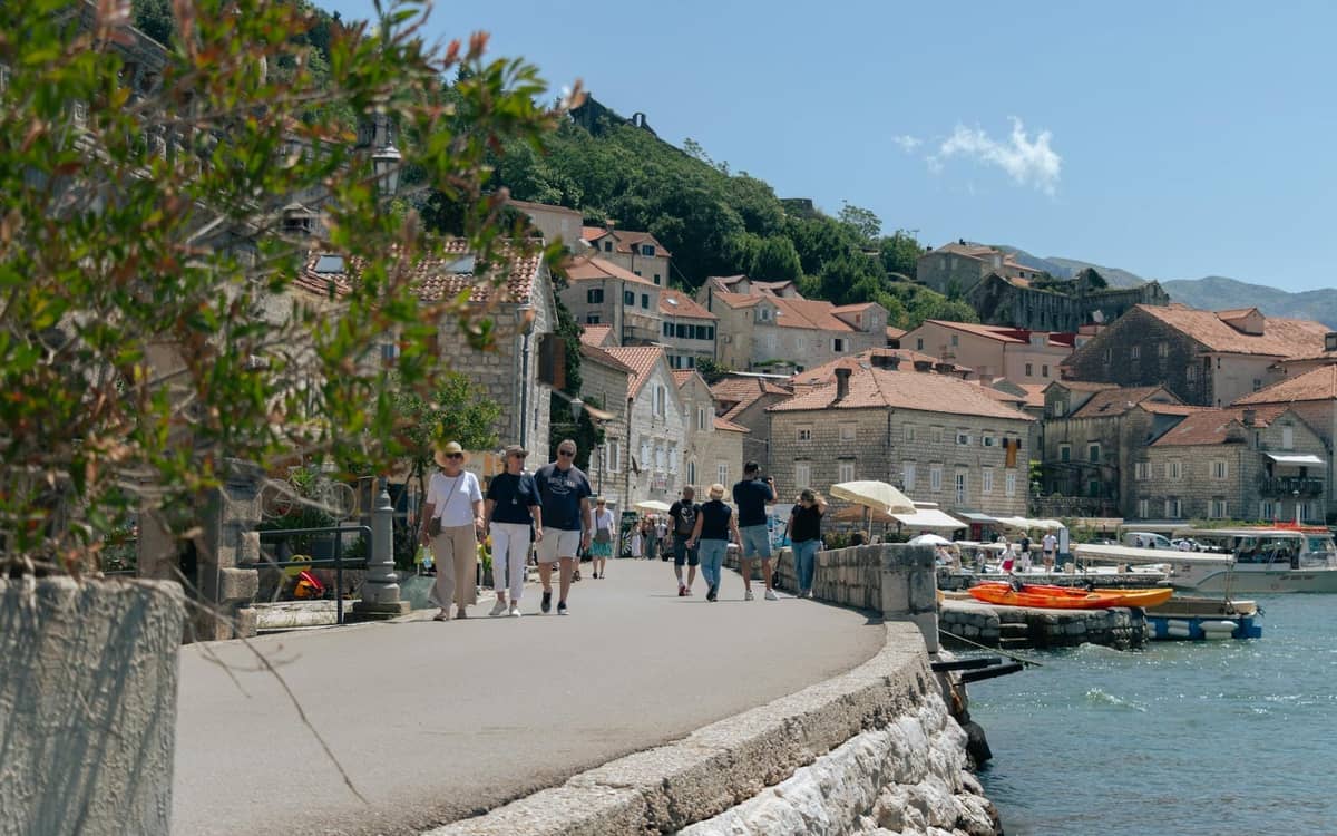 People walking along Kotor’s waterfront promenade with stone buildings, small boats, and bay waters at ground level on a sunny day.