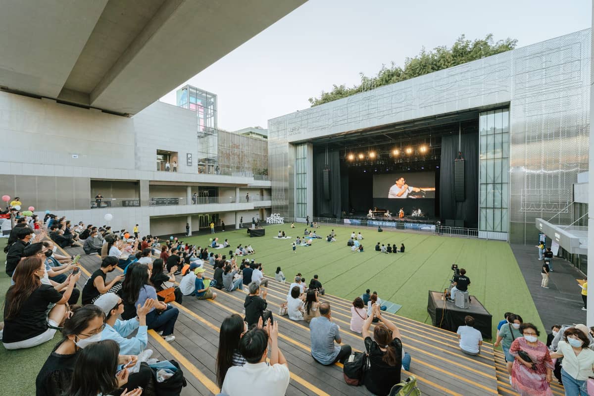 Público ao ar livre sentado em degraus em frente a um palco moderno, assistindo à gravação de um show ao vivo em um espaço de apresentações em Seoul.