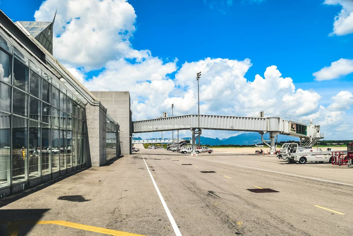 Vista exterior do Aeroporto Internacional de Krabi com janelas de vidro, ponte de embarque, pista e montanhas ao fundo sob céu azul com nuvens