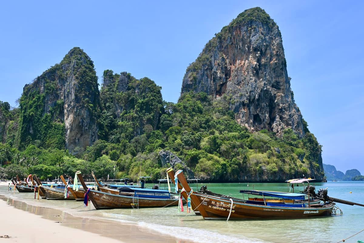 Barcos de cauda longa na orla arenosa com água turquesa cristalina e dramáticos penhascos de calcário em Railay Beach, Krabi, Tailândia.