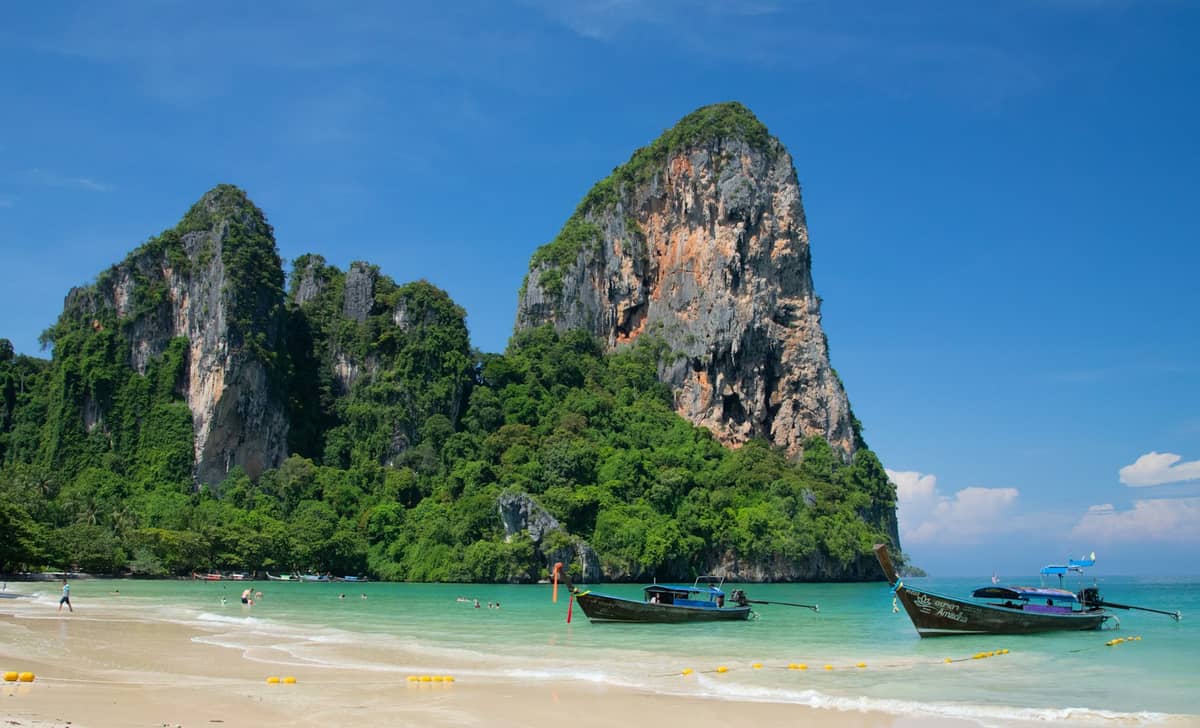 Vue panoramique de la plage de Railay à Krabi, Thaïlande, avec ses imposantes falaises calcaires, ses eaux turquoise et ses bateaux longtail ancrés près du rivage sous un ciel bleu.