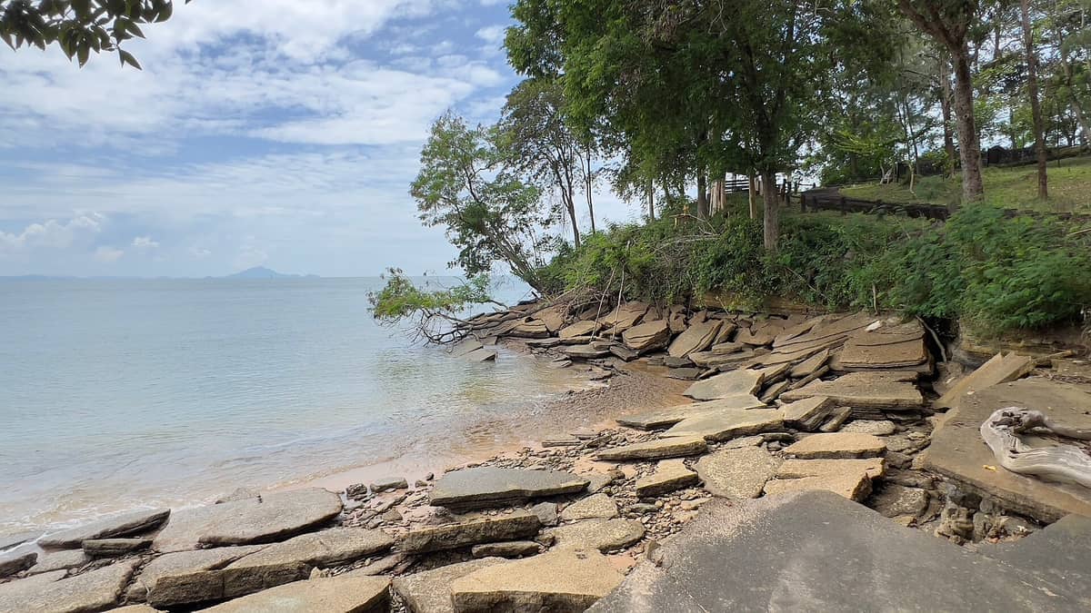 Große, flache Fossilgesteine sind entlang des Ufers bei Susan Hoi, Fossil Shell Beach, verstreut, mit ruhigem Meerwasser und üppigen Bäumen unter einem teilweise bewölkten Himmel.