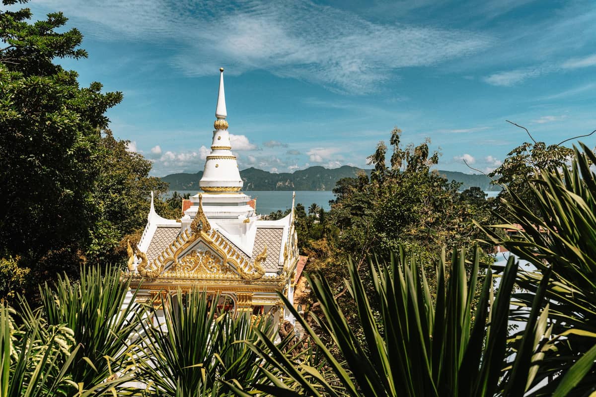 Verzierter thailändischer Tempel mit goldenem Dach und weißer Stupa, umgeben von üppigem Grün, mit Blick auf Ozean und Berge im Hintergrund unter blauem Himmel