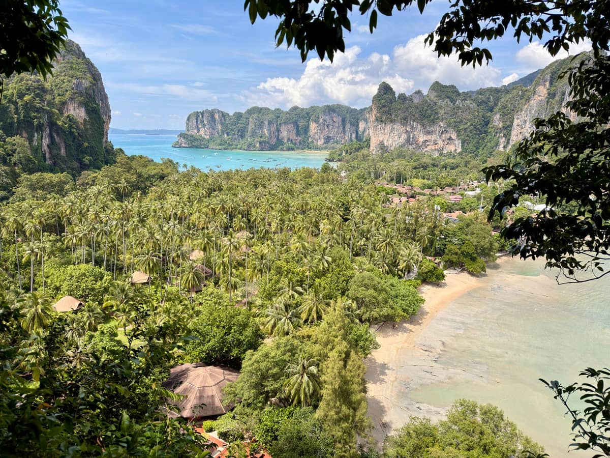 Panoramablick auf den Railay Beach in Krabi, Thailand, mit üppigem Dschungel, Palmen, dramatischen Kalksteinklippen und türkisblauem Wasser in der Ferne.