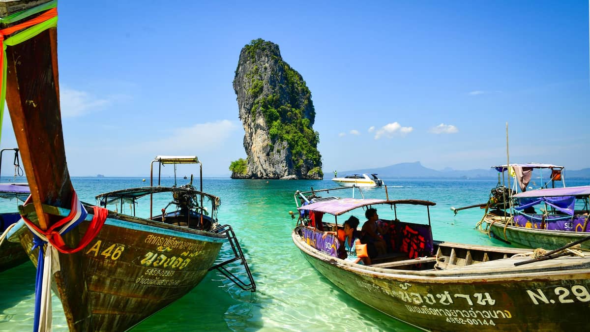 Traditionelle thailändische Longtail-Boote treiben in klarem türkisfarbenem Wasser mit dramatischer Kalksteininselszenerie in Krabi, Thailand, unter einem strahlend blauen Himmel.