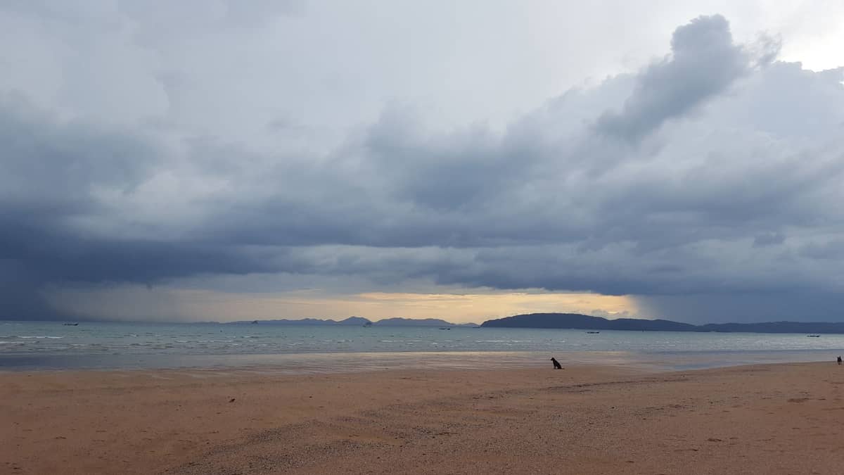 Vaste plage de Krabi avec de sombres nuages de mousson et des îles lointaines sur une eau calme, illustrant l'atmosphère de la saison des pluies.