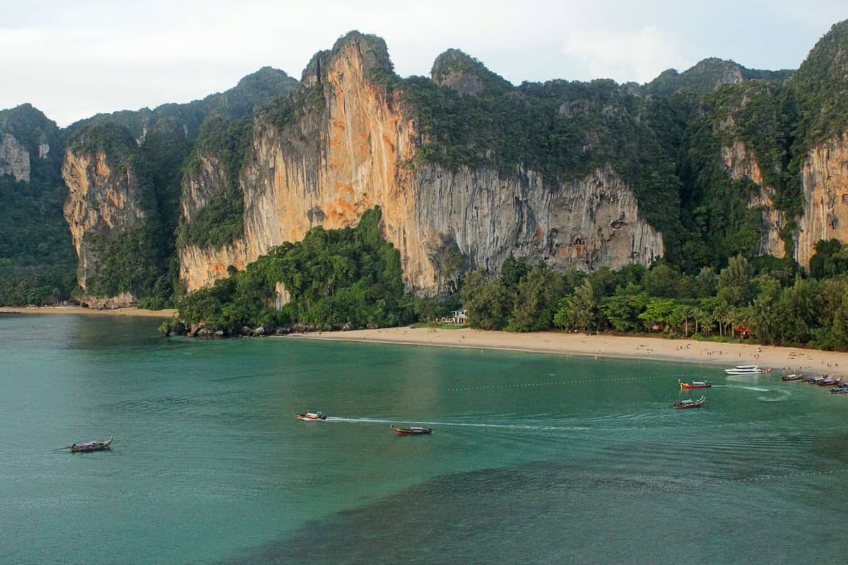 Vista aérea de la costa de Krabi con imponentes acantilados de piedra caliza, playa de arena, agua turquesa y varios barcos de cola larga en alta mar.