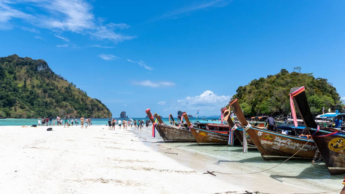 Varios barcos de cola larga tradicionales alineados en una playa de arena blanca con turistas caminando por un banco de arena entre exuberantes islas, bajo un cielo azul brillante