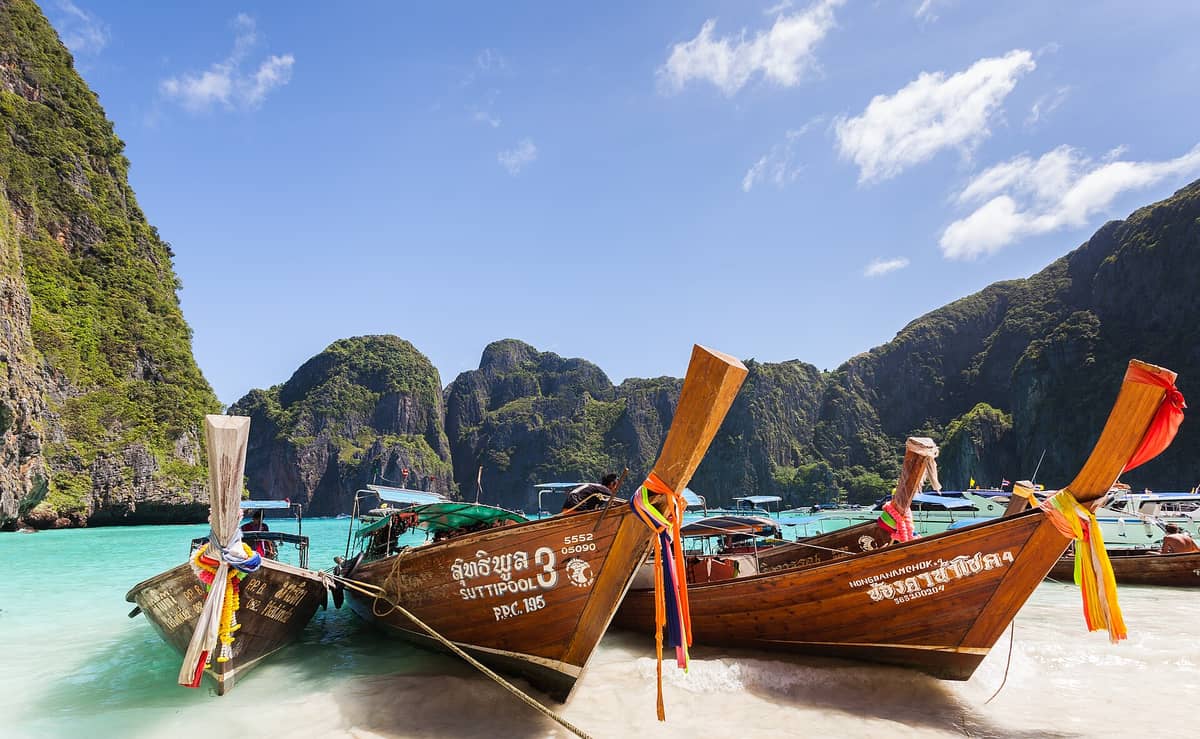 Barcas tradicionales de cola larga ancladas en la playa de Maya Bay, con imponentes acantilados de piedra caliza y agua turquesa bajo un cielo despejado.
