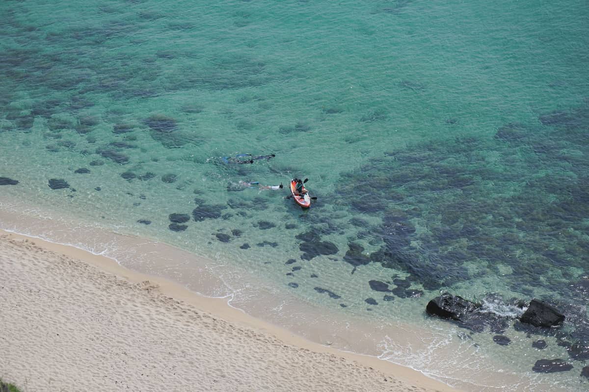 Vista aérea de una persona en kayak cerca de la playa en aguas turquesas cristalinas con esnorquelistas visibles cerca.