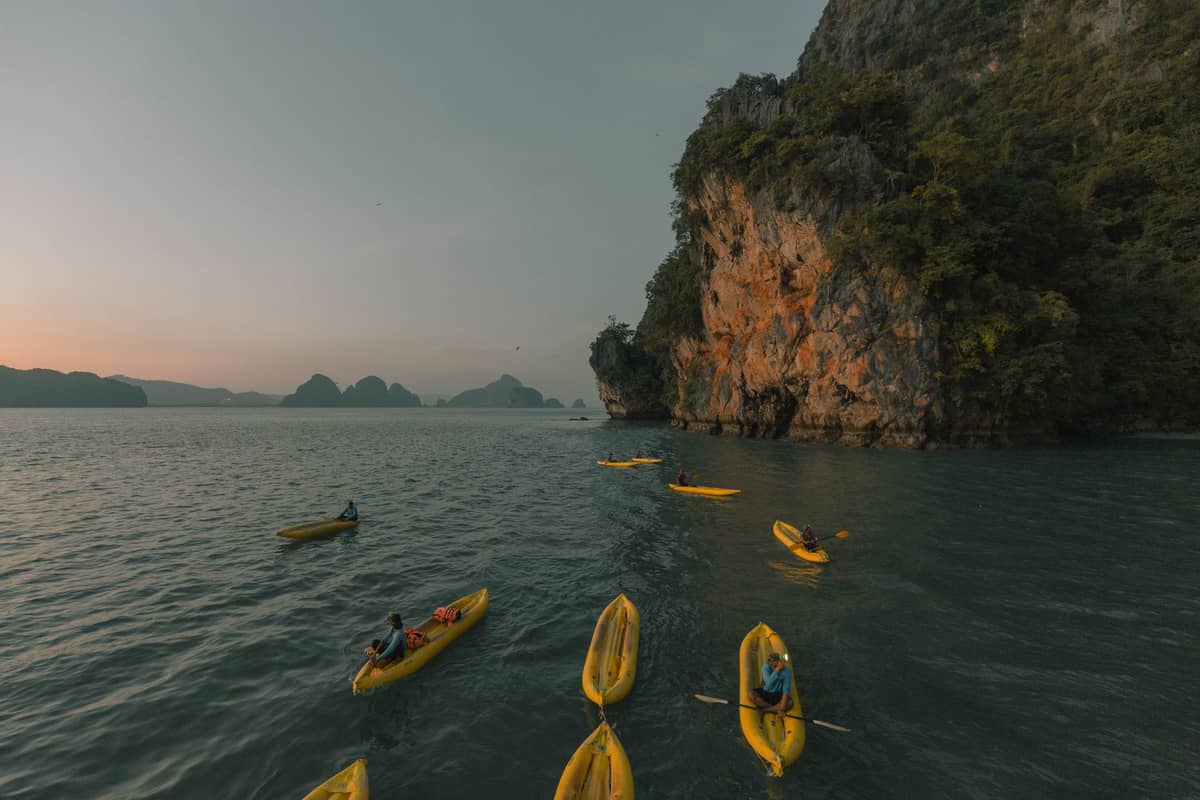 Varios kayaks amarillos navegan junto a imponentes acantilados de piedra caliza y aguas tranquilas al atardecer en Krabi, Tailandia, con islas distantes en el horizonte.
