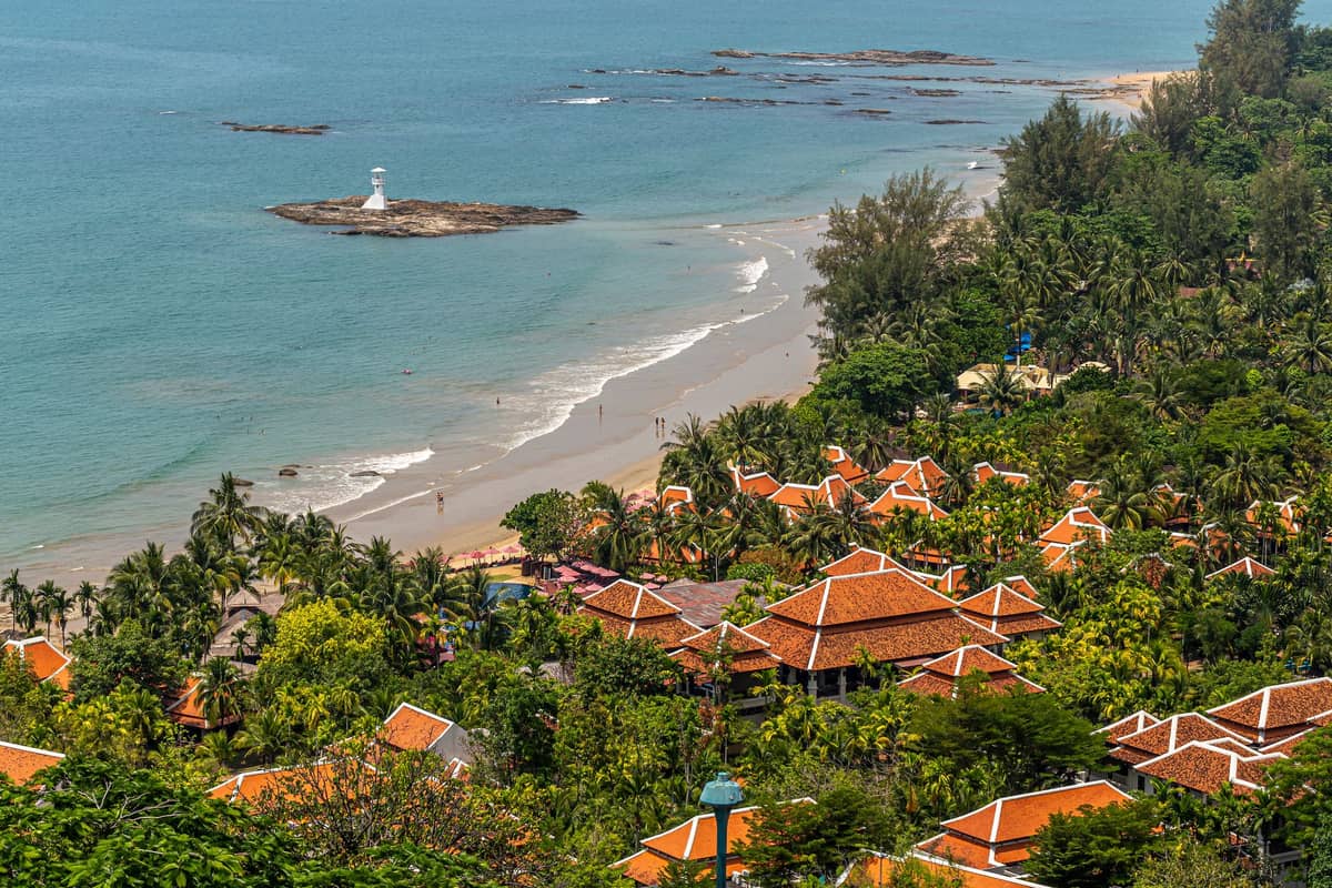 Vue aérienne du littoral de Koh Lanta avec une plage de sable, des hébergements aux toits rouges, des palmiers et un petit phare sur un îlot rocheux au large.