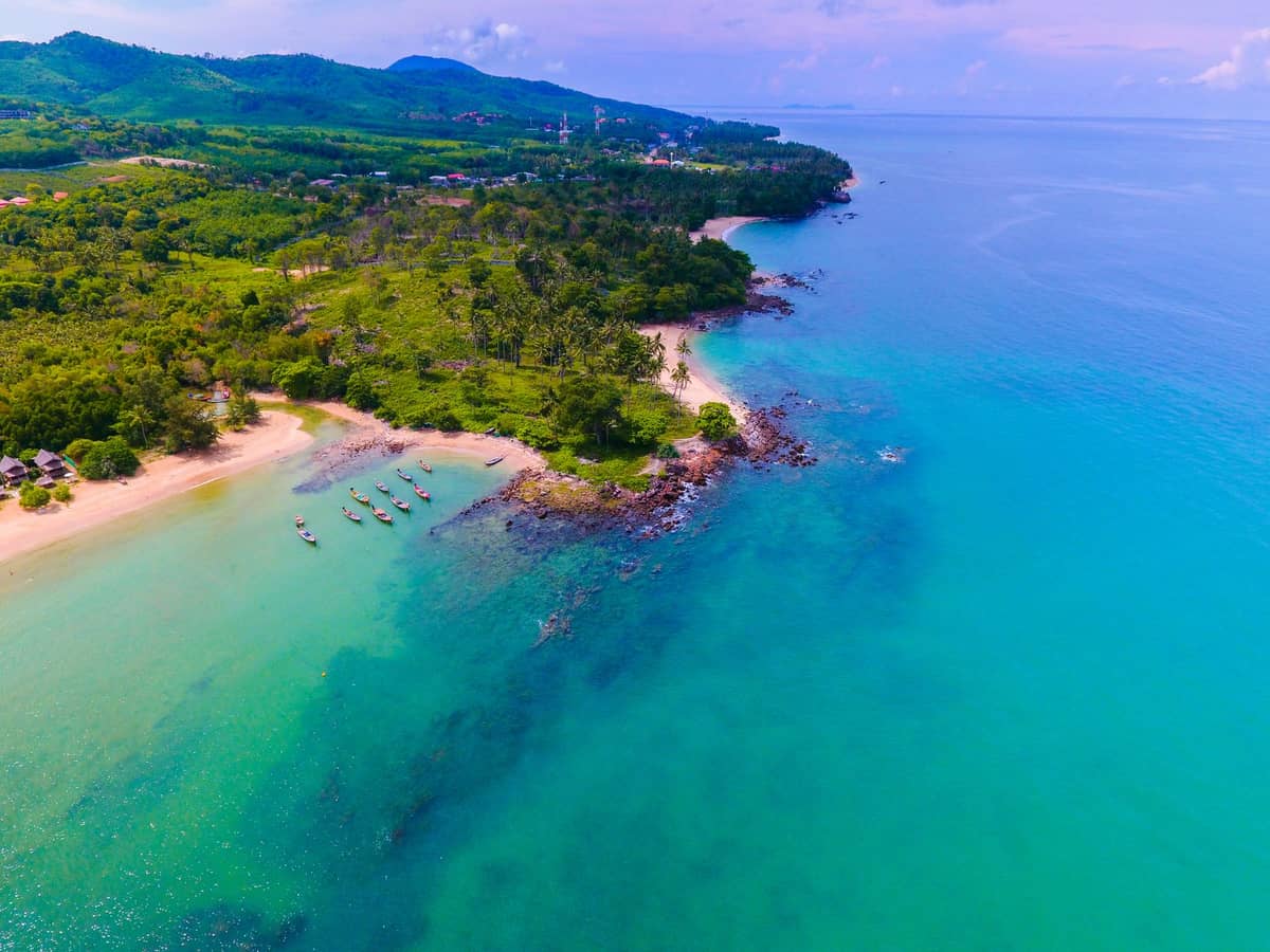 Vue aérienne du littoral de Koh Lanta avec une eau turquoise, des plages de sable, de petits bateaux, des collines verdoyantes et un panorama côtier sous un ciel partiellement nuageux.
