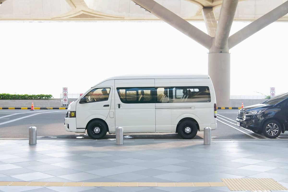 Minivan blanche garée en bordure de trottoir dans un aéroport, avec un sol carrelé, des bollards et une partie d'un autre véhicule noir visible à proximité.