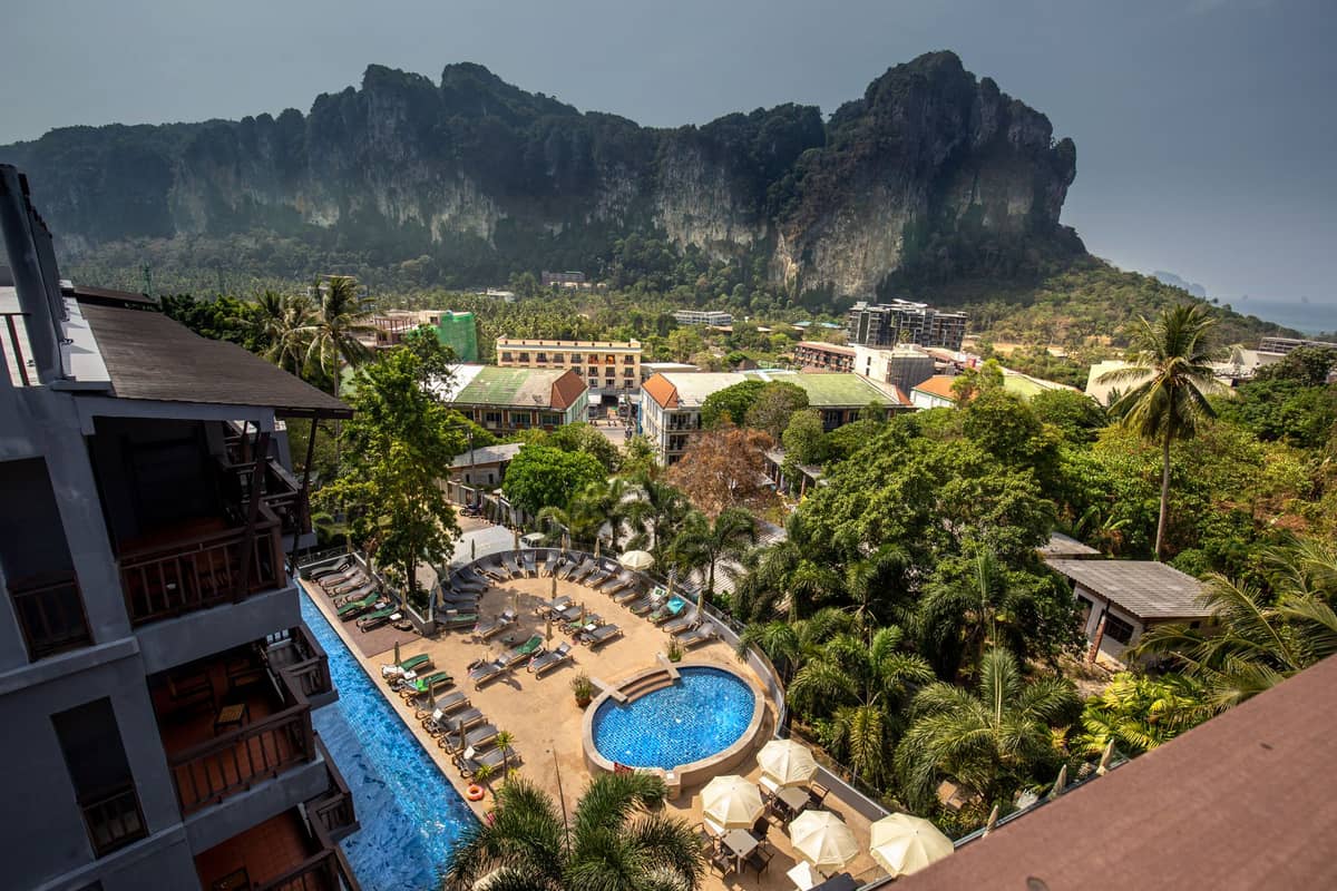 Blick auf den Poolbereich eines Luxusresorts mit Sonnenliegen, Palmen und beeindruckenden Kalksteinfelsen im Hintergrund, typisch für Krabi, Thailand.