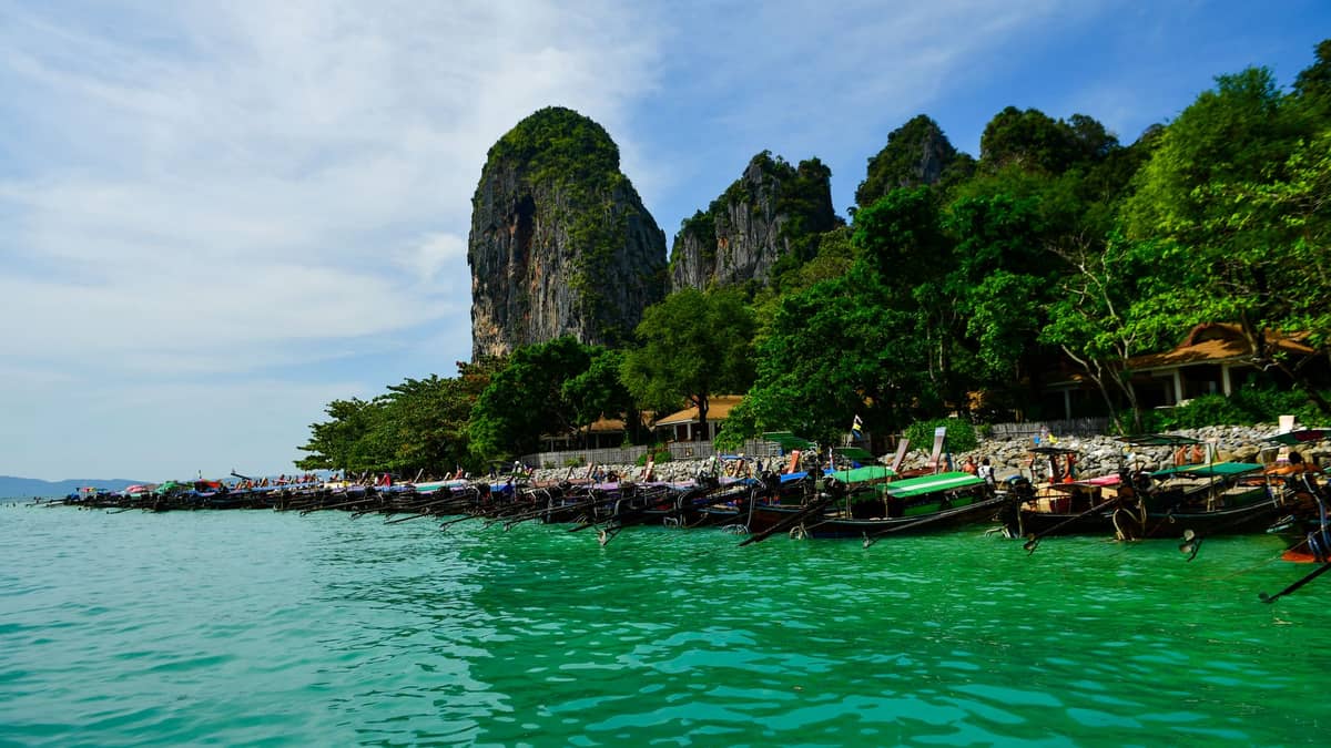 Łodzie longtail ustawione wzdłuż turkusowego brzegu Railay Beach z dramatycznymi klifami wapiennymi i bujną zielenią w tle.