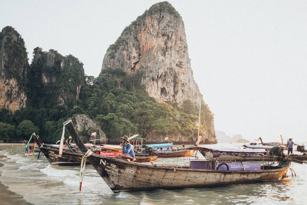 Des bateaux à longue queue flottent près du rivage, avec les spectaculaires falaises calcaires qui s'élèvent derrière eux — la vue emblématique de la plage de Railay à Krabi, en Thaïlande.