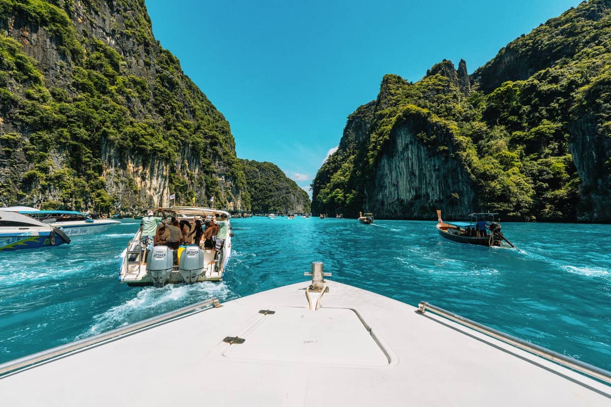 Blick vom Bug eines Schnellboots, das sich einer türkisfarbenen Lagune mit Klippen, Touristenbooten und Longtails bei den Phi Phi Islands, Thailand, nähert.
