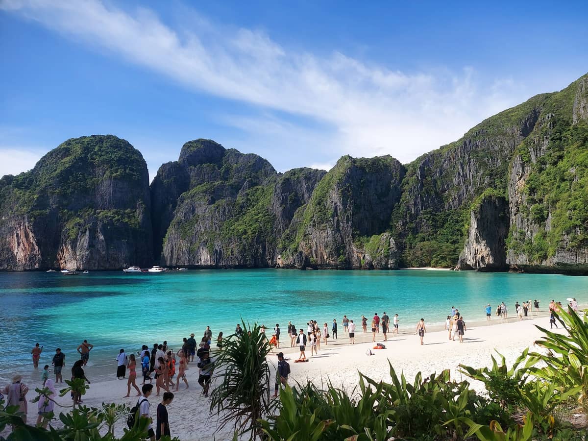 Weiter Blick auf die Maya Bay auf Koh Phi Phi mit türkisblauem Wasser, dramatischen Kalksteinfelsen und Besuchern, die unter blauem Himmel am Sandstrand entlanglaufen.