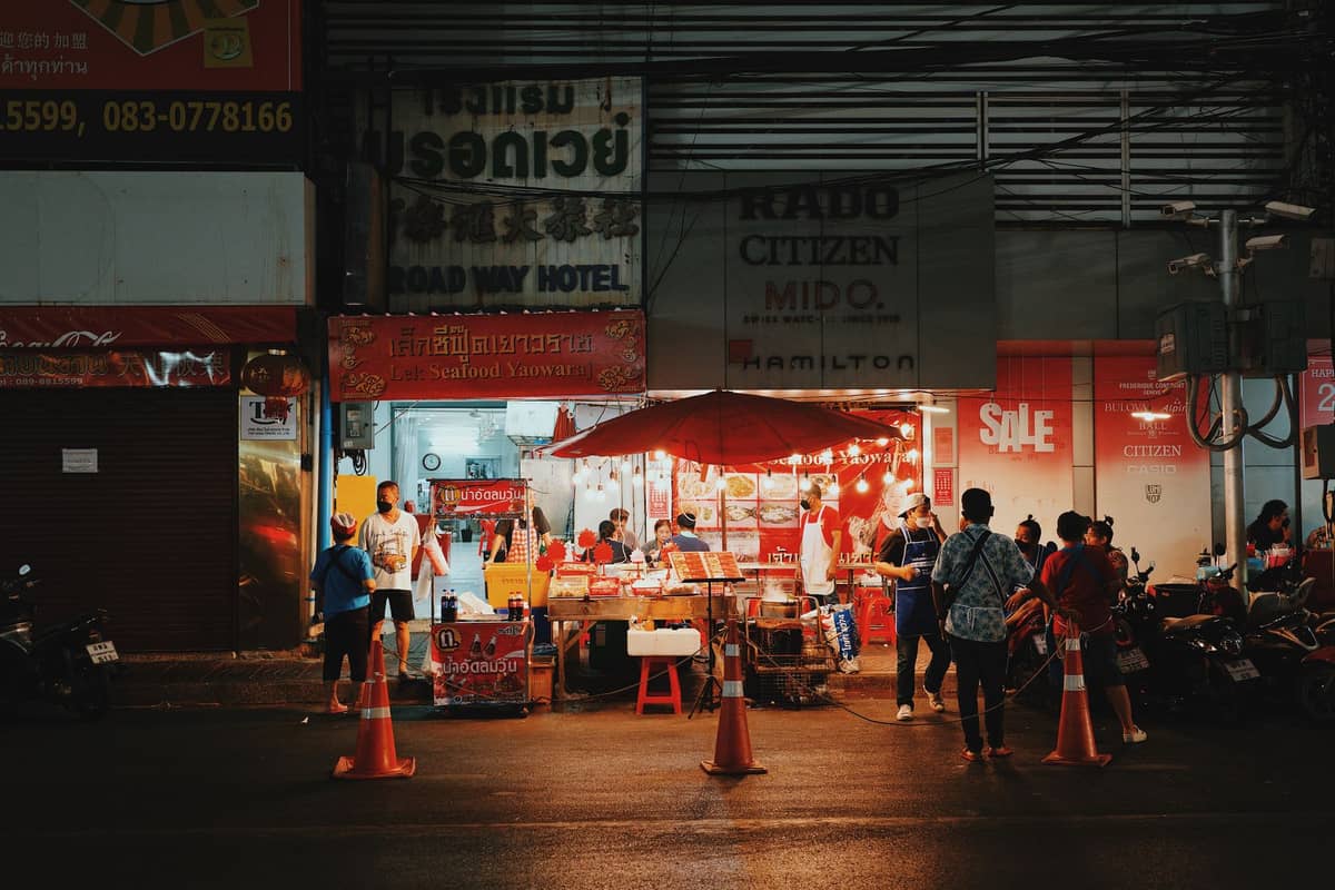 Straßenimbissverkäufer unter Schirmen und hellen Lichtern auf einer belebten thailändischen Straße bei Nacht mit Menschen, die Schlange stehen und einkaufen.