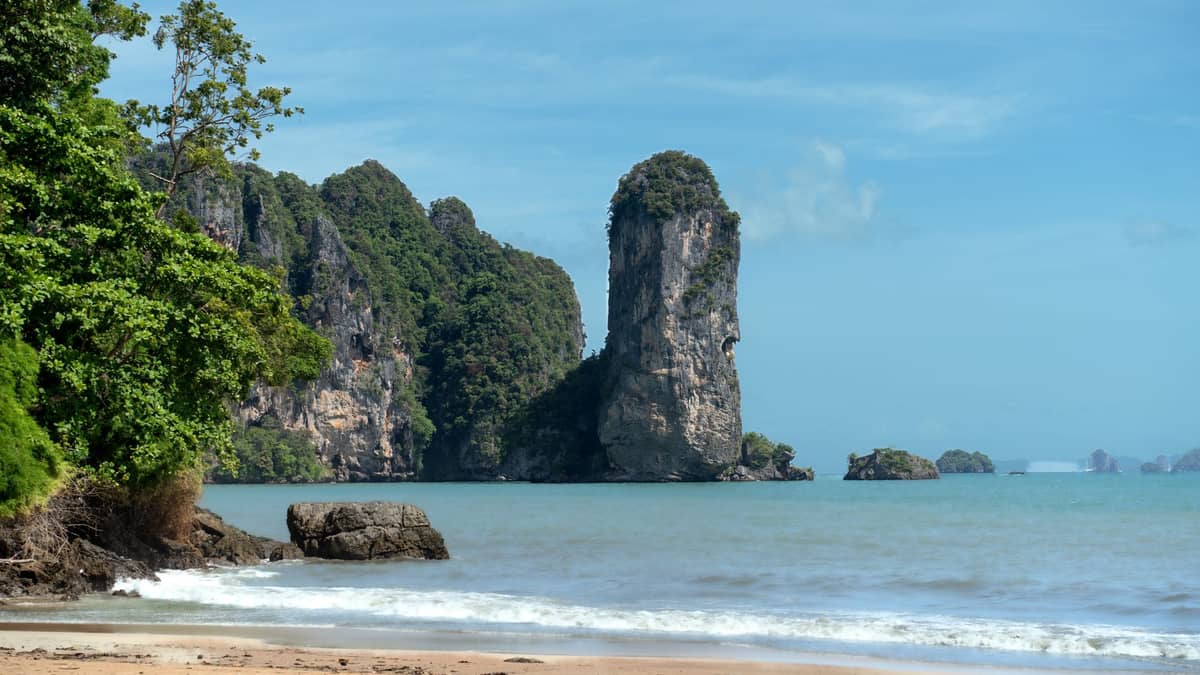 Coastal view of Krabi, Thailand with a distinctive limestone karst jutting out from turquoise waters, lush green cliffs, and a sandy tropical beach under blue sky.