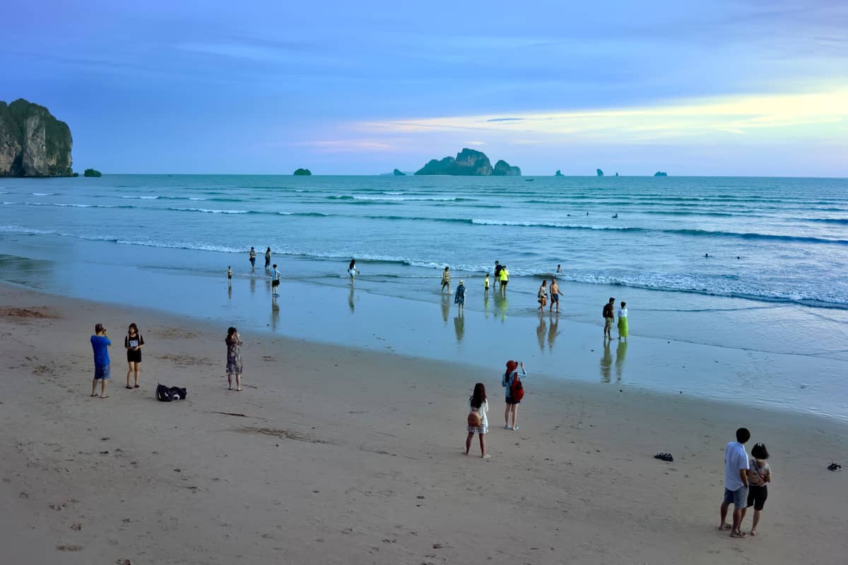 Menschen verteilt am Ao Nang Beach in der Abenddämmerung; flacher brauner Sand, sanfte Wellen und die typischen Kalksteinfelsen von Krabi im Hintergrund.
